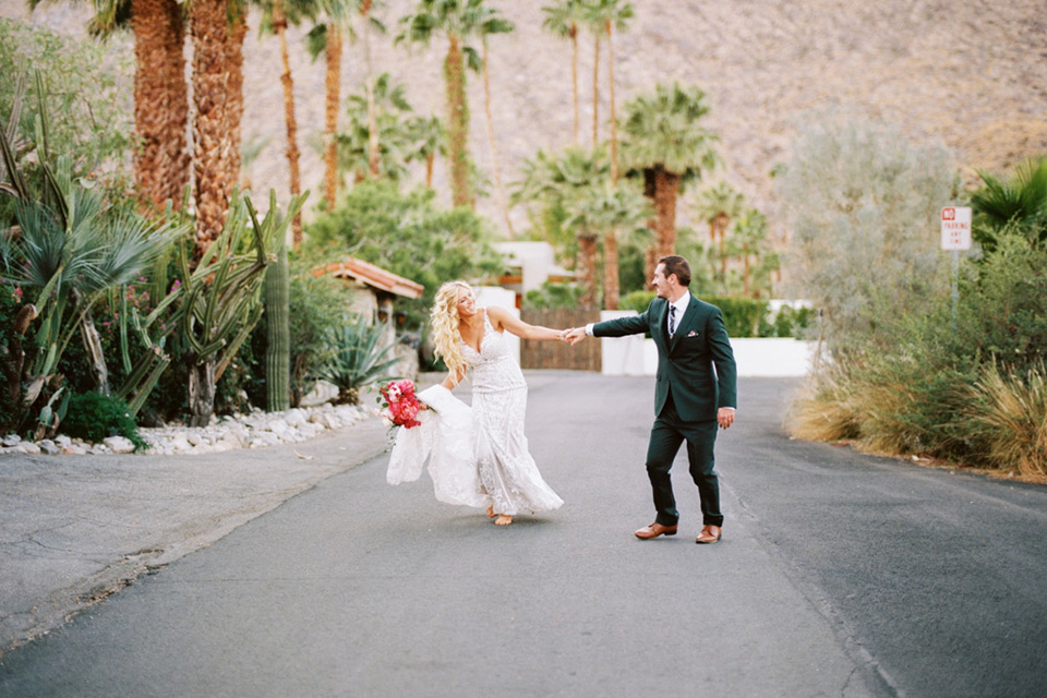  bride in a vintage bohemian gown with beaded details and cape, groom in a green suit with a floral tie sitting on couch