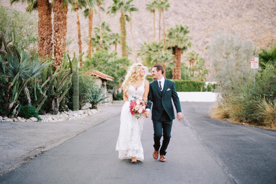  bride in a vintage bohemian gown with beaded details and cape, groom in a green suit with a floral tie sitting on couch