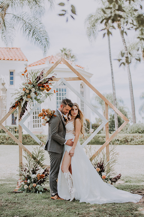  bride in a bohemian lace gown with a tulle skirt, lace boots, and headpiece and the groom in a café brown suit with an ivory tie, with a geometric arch 