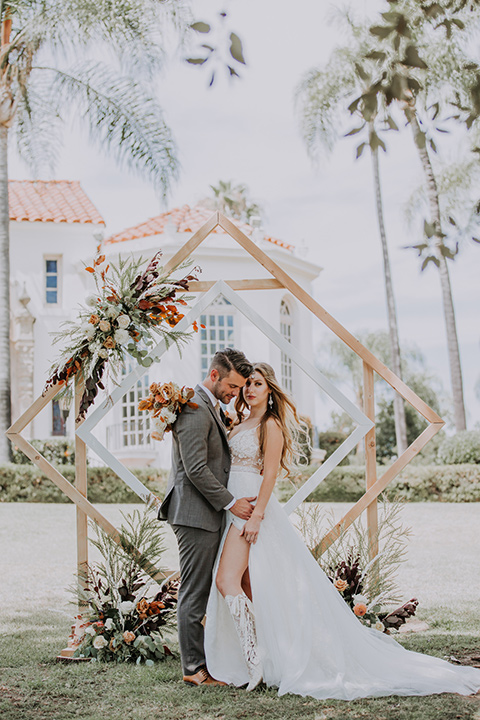  bride in a bohemian lace gown with a tulle skirt, lace boots, and headpiece and the groom in a café brown suit with an ivory tie, with a geometric arch