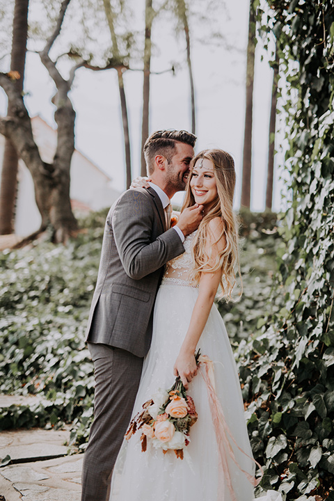  bride in a bohemian lace gown with a tulle skirt, lace boots, and headpiece and the groom in a café brown suit with an ivory tie, with a geometric arch