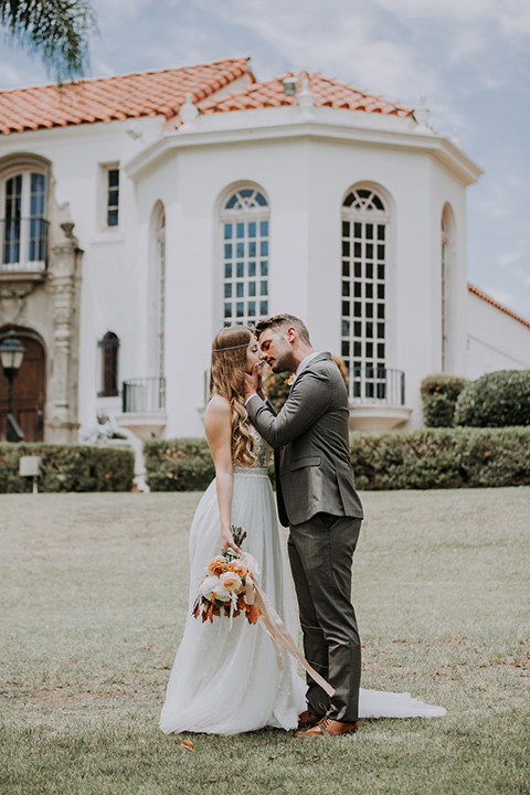  bride in a bohemian lace gown with a tulle skirt, lace boots, and headpiece and the groom in a café brown suit with an ivory tie, with a geometric arch