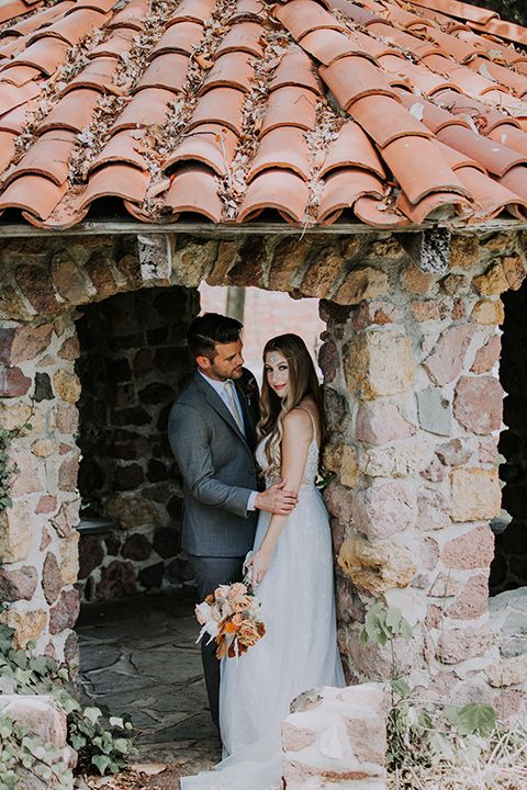  bride in a bohemian lace gown with a tulle skirt, lace boots, and headpiece and the groom in a café brown suit with an ivory tie, with a geometric arch 