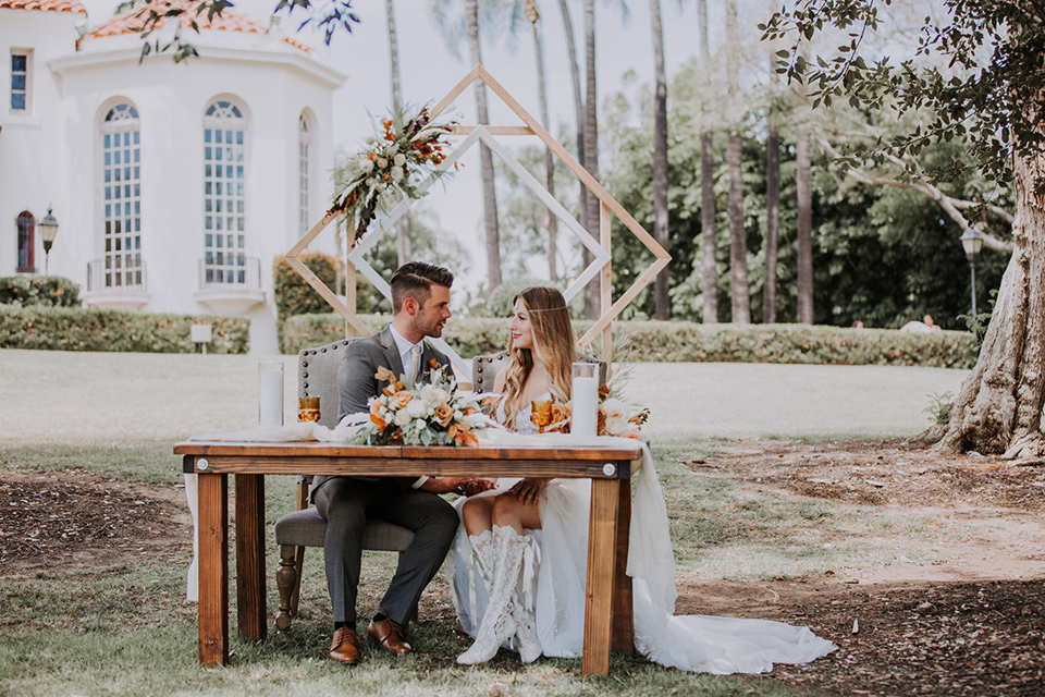  bride in a bohemian lace gown with a tulle skirt, lace boots, and headpiece and the groom in a café brown suit with an ivory tie sitting at a sweetheart table