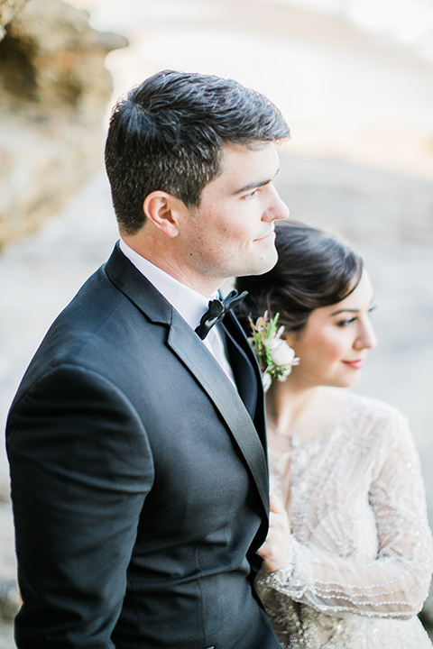 bride in a flowing gown with an illusion neckline and hair in a soft updo with a crystal pin detail and the groom in a black notch lapel tuxedo with a black bow tie holding his bride close