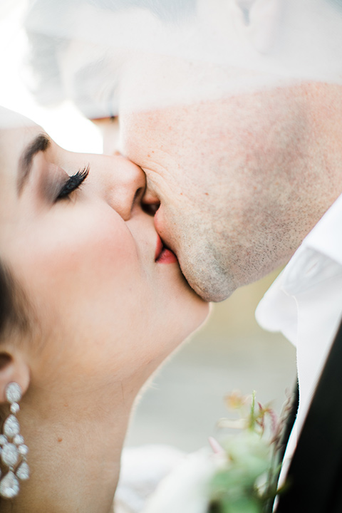 bride in a flowing gown with an illusion neckline and hair in a soft updo with a crystal pin detail and the groom in a black notch lapel tuxedo with a black bow tie kissing