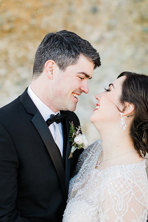 bride in a flowing gown with an illusion neckline and hair in a soft updo with a crystal pin detail and the groom in a black notch lapel tuxedo with a black bow tie kissing