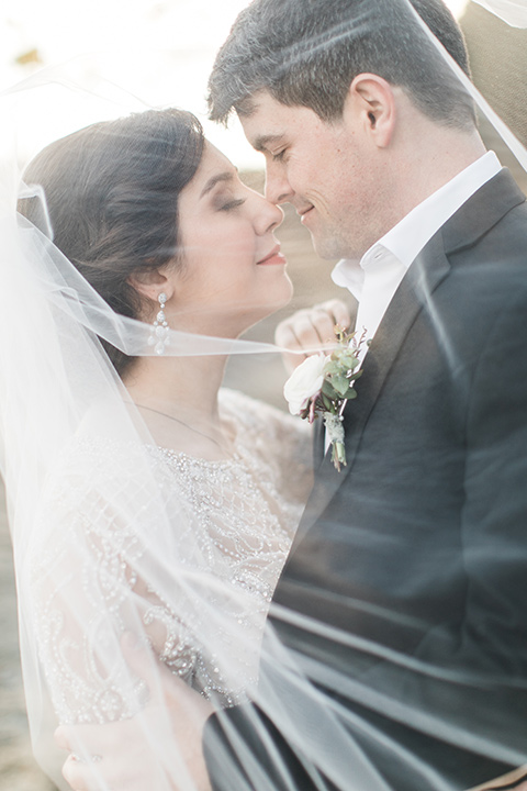  bride in a flowing gown with an illusion neckline and hair in a soft updo with a crystal pin detail and the groom in a black notch lapel suit with a black bow tie under veil 