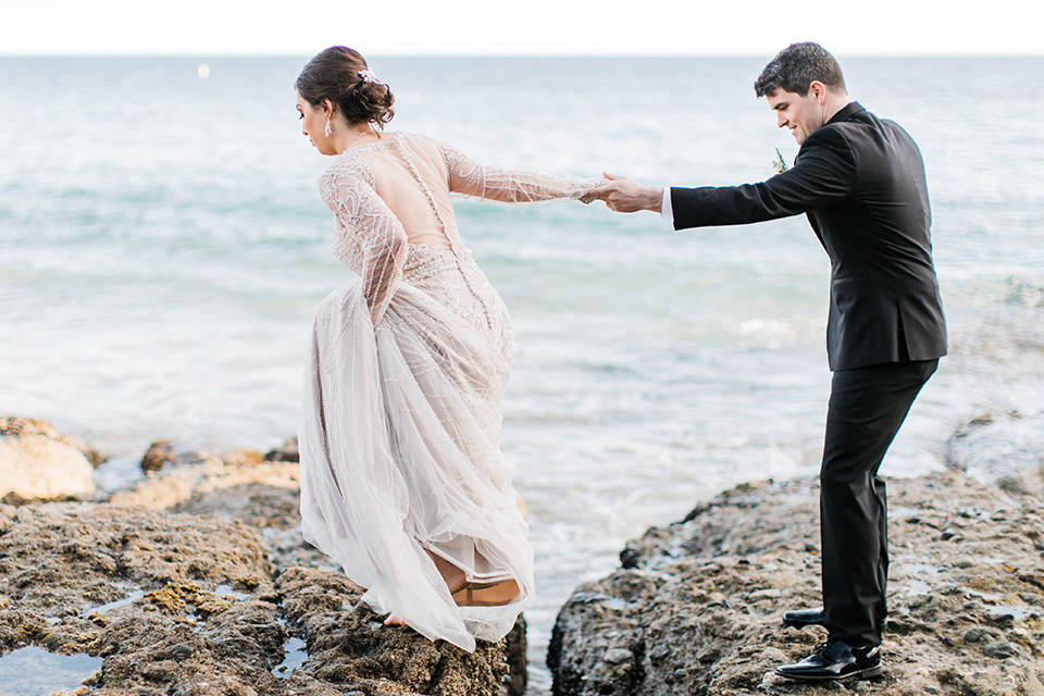  bride in a flowing gown with an illusion neckline and hair in a soft updo with a crystal pin detail and the groom in a black notch lapel tuxedo with a black bow tie holding hands