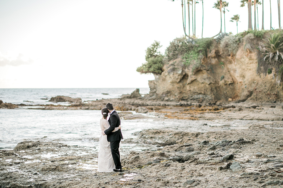  bride in a flowing gown with an illusion neckline and hair in a soft updo with a crystal pin detail and the groom in a black notch lapel tuxedo with a black bow tie on the rocks