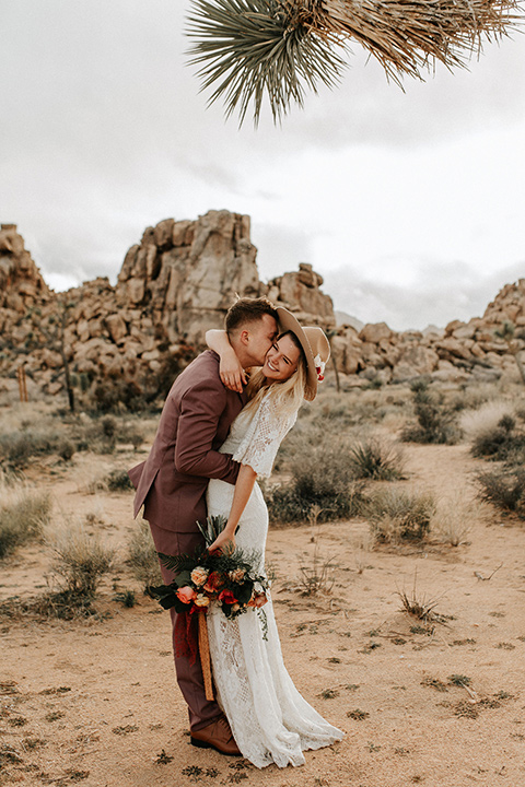  bride in a lace bohemian gown with flowing sleeves and wide brimmed hat and the groom in a rose pink suit with a tan long tie 