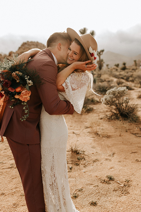  bride in a lace bohemian gown with flowing sleeves and wide brimmed hat and the groom in a rose pink suit with a tan long tie 