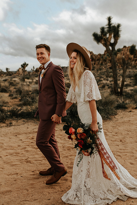  bride in a lace bohemian gown with flowing sleeves and wide brimmed hat and the groom in a rose pink suit with a tan long tie 