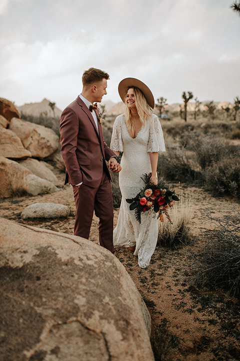  bride in a lace bohemian gown with flowing sleeves and wide brimmed hat and the groom in a rose pink suit with a tan long tie 