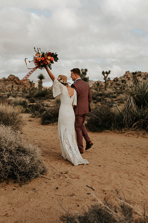  bride in a lace bohemian gown with flowing sleeves and wide brimmed hat and the groom in a rose pink suit with a tan long tie 