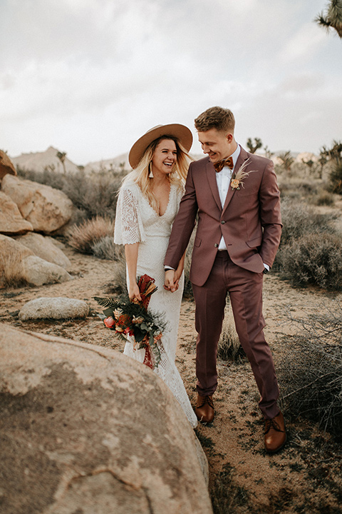  bride in a lace bohemian gown with flowing sleeves and wide brimmed hat and the groom in a rose pink suit with a tan long tie 