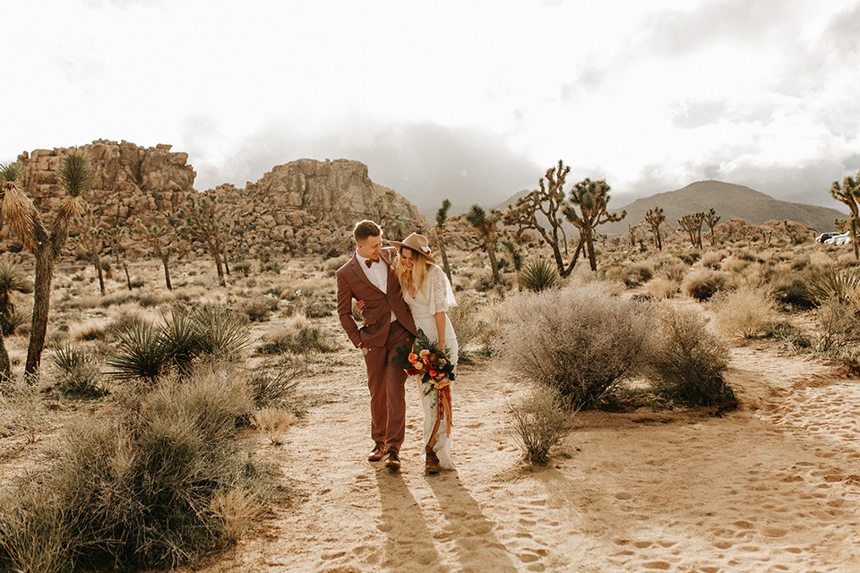  bride in a lace bohemian gown with flowing sleeves and wide brimmed hat and the groom in a rose pink suit with a tan long tie 