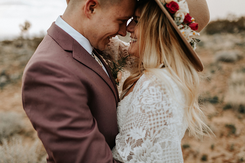  bride in a lace bohemian gown with flowing sleeves and wide brimmed hat and the groom in a rose pink suit with a tan long tie 