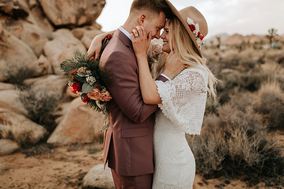  bride in a lace bohemian gown with flowing sleeves and wide brimmed hat and the groom in a rose pink suit with a tan long tie 