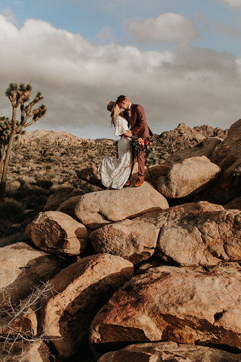  bride in a lace bohemian gown with flowing sleeves and wide brimmed hat and the groom in a rose pink suit with a tan long tie 