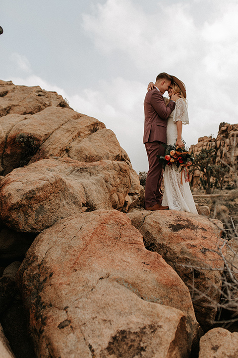  bride in a lace bohemian gown with flowing sleeves and wide brimmed hat and the groom in a rose pink suit with a tan long tie 