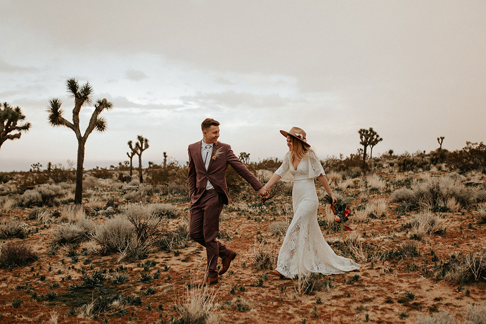  bride in a lace bohemian gown with flowing sleeves and wide brimmed hat and the groom in a rose pink suit with a tan long tie 