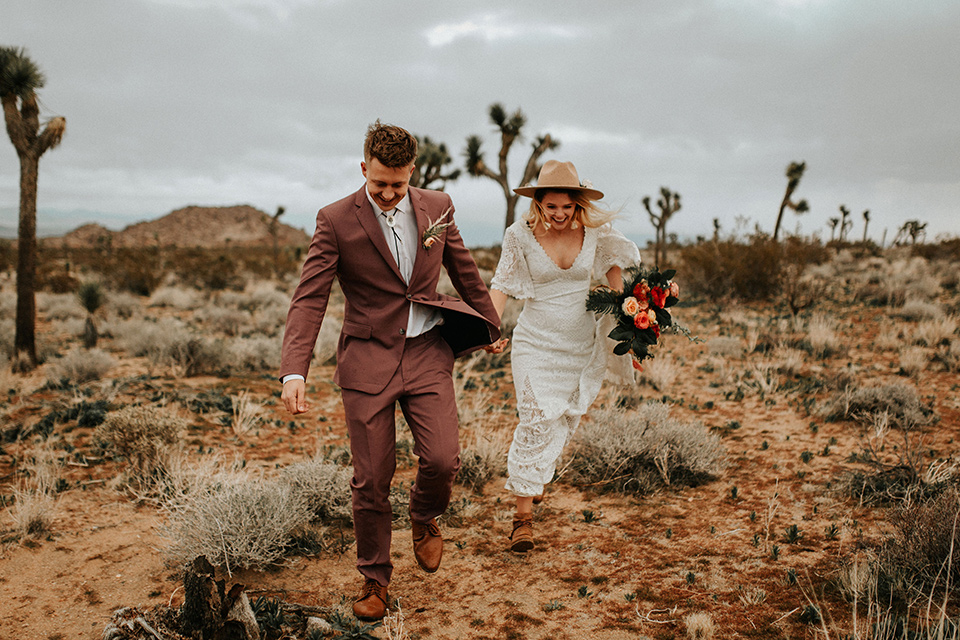  bride in a lace bohemian gown with flowing sleeves and wide brimmed hat and the groom in a rose pink suit with a tan long tie 