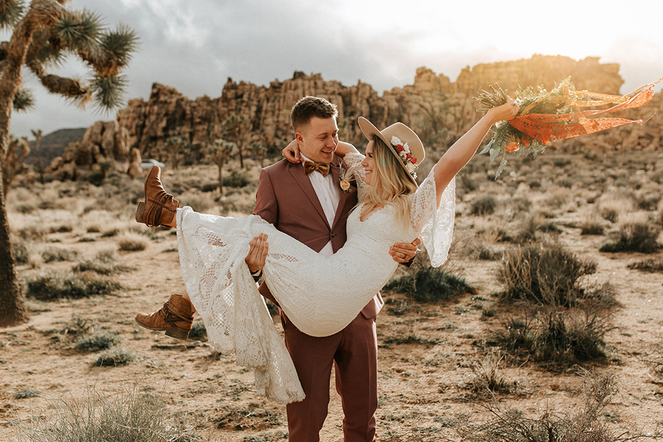  bride in a lace bohemian gown with flowing sleeves and wide brimmed hat and the groom in a rose pink suit with a tan long tie 