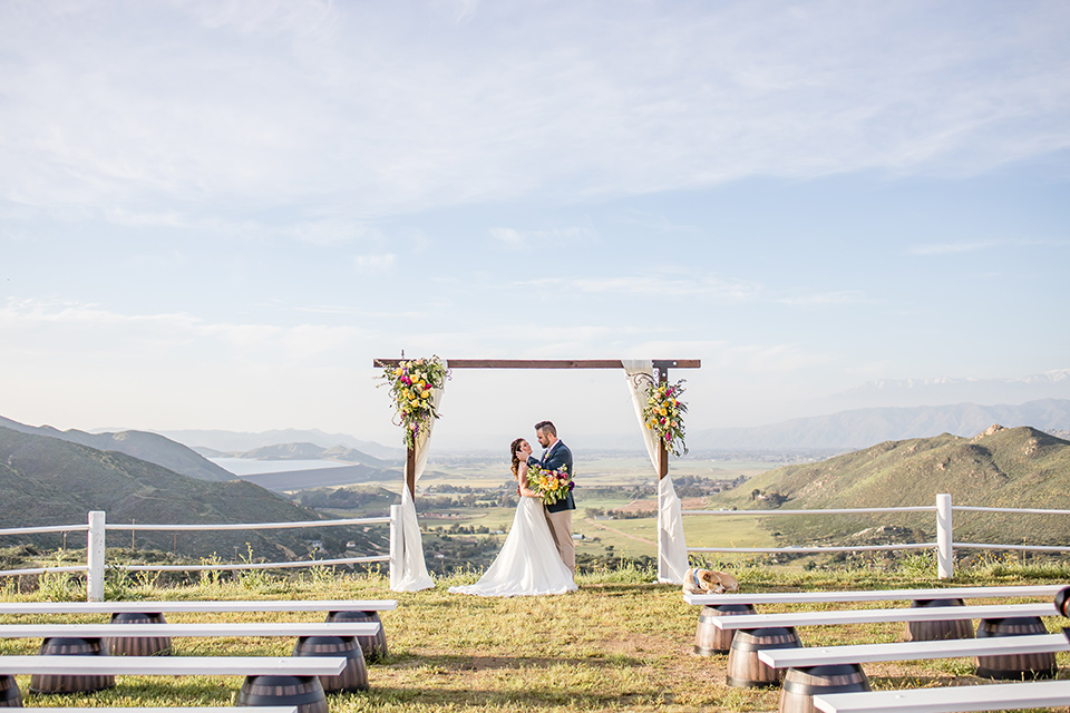  bride in a lace gown with thin straps and beaded bodice, the groom in a blue suit coat and tan pants with a white long tie 