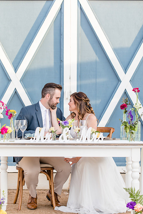  bride in a lace gown with thin straps and beaded bodice, the groom in a blue suit coat and tan pants with a white long tie, sitting at the table 