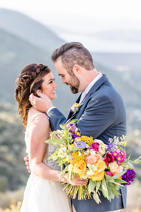  bride in a lace gown with thin straps and beaded bodice, the groom in a blue suit coat and tan pants with a white long tie close together 