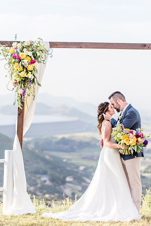  bride in a lace gown with thin straps and beaded bodice, the groom in a blue suit coat and tan pants with a white long tie, sitting at the table 