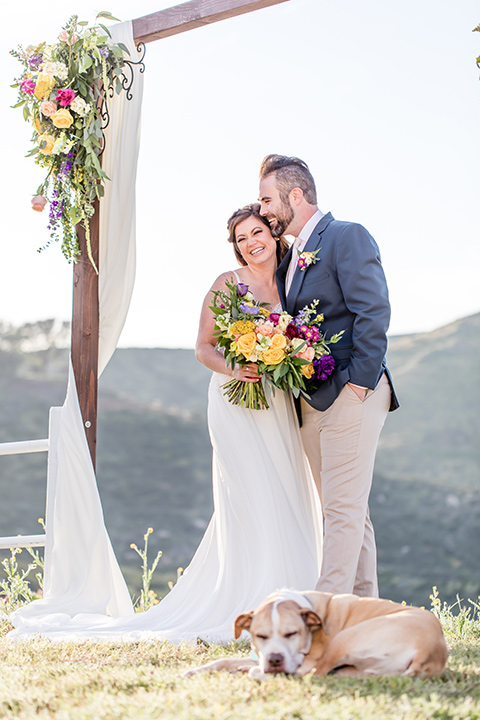  bride in a lace gown with thin straps and beaded bodice, the groom in a blue suit coat and tan pants with a white long tie, sitting at the table 