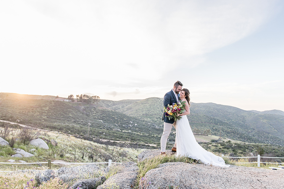  bride in a lace gown with thin straps and beaded bodice, the groom in a blue suit coat and tan pants with a white long tie 