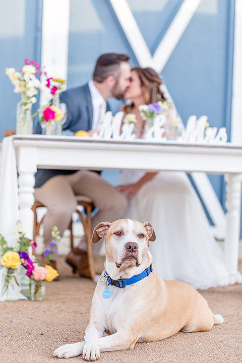  bride in a lace gown with thin straps and beaded bodice, the groom in a blue suit coat and tan pants with a white long tie, sitting at the table 