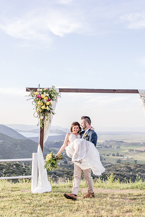  bride in a lace gown with thin straps and beaded bodice, the groom in a blue suit coat and tan pants with a white long tie close together 