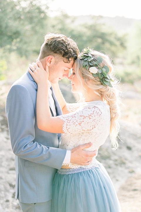 bride in a two-piece gown with a blue tulle skirt and white lace shirt and her hair in a loose French braid and floral crown the groom in a light blue suit with no tie and brown shoes