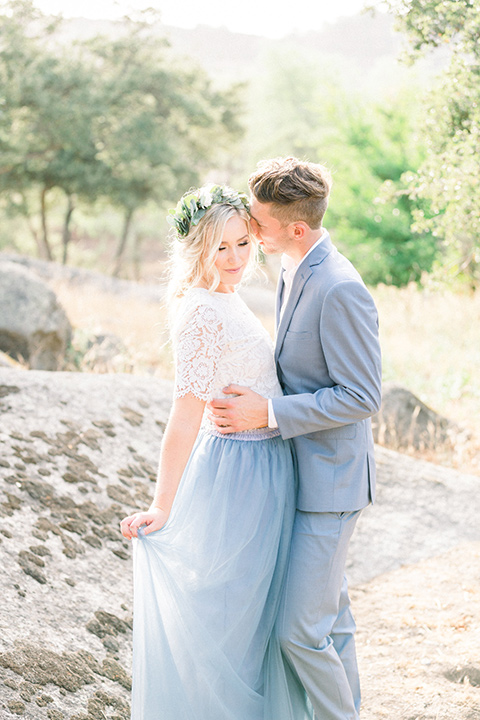  bride in a two-piece gown with a blue tulle skirt and white lace shirt and her hair in a loose French braid and floral crown the groom in a light blue suit with no tie and brown shoes