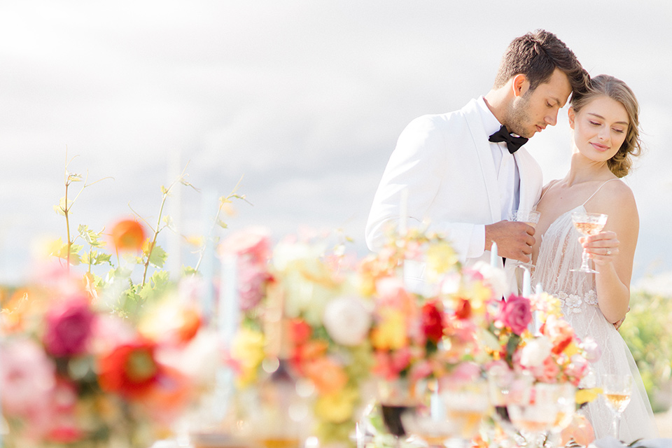  bride in a white flowing gown with a v neckline and the groom in a white dinner jacket and black pants 