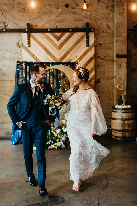  bride in an ivory lace gown with flowing sleeves and a braided hairstyle and the groom in a cobalt blue suit and a black long tie walking down the altar 