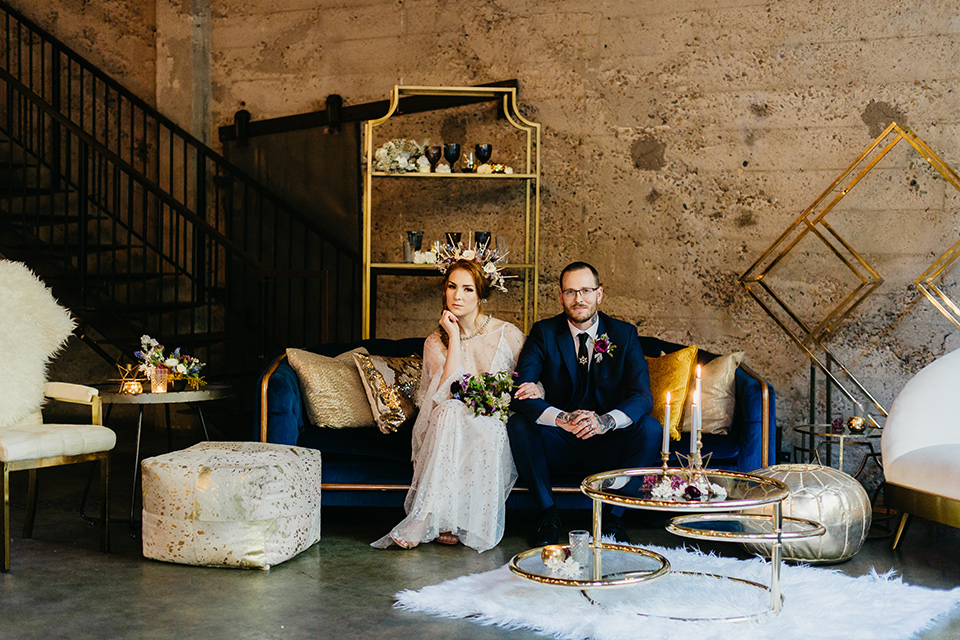  bride in an ivory lace gown with flowing sleeves and a braided hairstyle and the groom in a cobalt blue suit and a black long tie on the couch