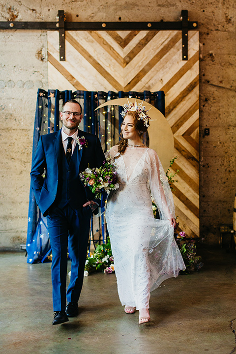  bride in an ivory lace gown with flowing sleeves and a braided hairstyle and the groom in a cobalt blue suit and a black long tie walking down the altar