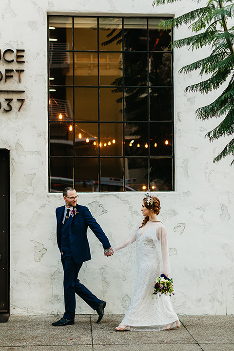  bride in an ivory lace gown with flowing sleeves and a braided hairstyle and the groom in a cobalt blue suit and a black long tie walking outside 