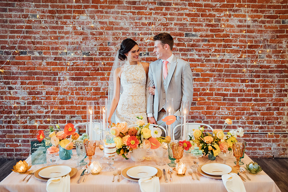  bride in a modern gown with a high neckline, no sleeves, and a tiered ruffled skirt and the groom in a light grey peak lapel suit with a coral long tie, by the sweetheart table