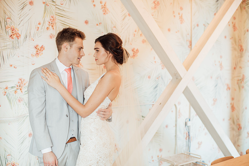  bride in a modern gown with a high neckline, no sleeves, and a tiered ruffled skirt and the groom in a light grey peak lapel suit with a coral long tie, in room with windows