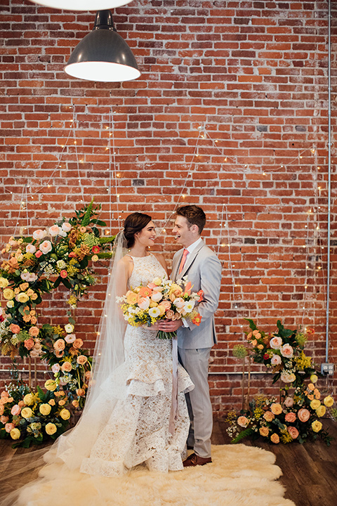  bride in a modern gown with a high neckline, no sleeves, and a tiered ruffled skirt and the groom in a light grey peak lapel suit with a coral long tie, at ceremony 