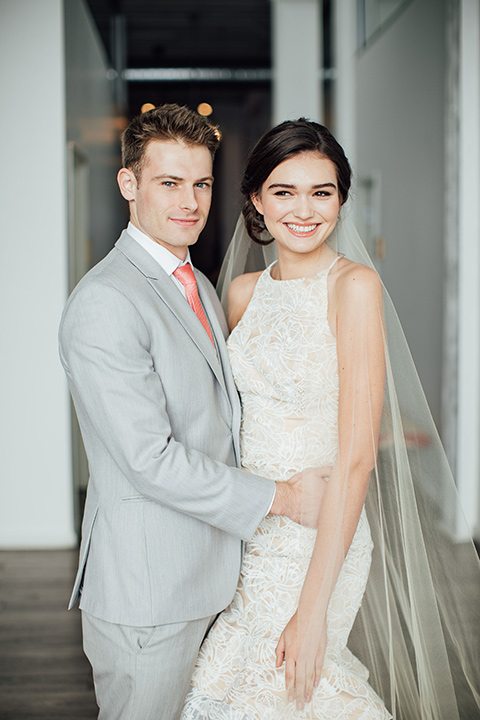  bride in a modern gown with a high neckline, no sleeves, and a tiered ruffled skirt and the groom in a light grey peak lapel suit with a coral long tie 