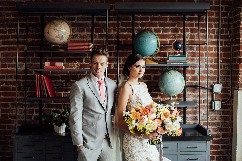  bride in a modern gown with a high neckline, no sleeves, and a tiered ruffled skirt and the groom in a light grey peak lapel suit with a coral long tie, in room with brick walls