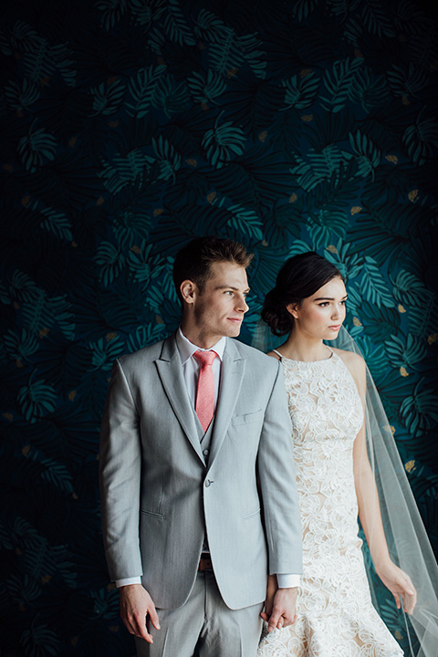  bride in a modern gown with a high neckline, no sleeves, and a tiered ruffled skirt and the groom in a light grey peak lapel suit with a coral long tie, at ceremony 