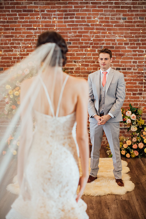  bride in a modern gown with a high neckline, no sleeves, and a tiered ruffled skirt and the groom in a light grey peak lapel suit with a coral long tie, at ceremony 